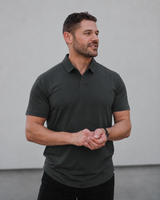 A man wearing a dark charcoal gray short-sleeve polo shirt and black pants, standing against a plain light gray wall.